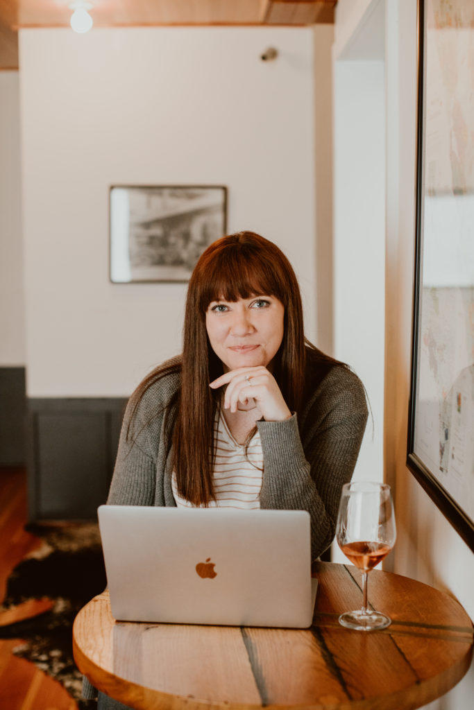 A woman sits at a table with a laptop and glass of wine in front of her while she smiles at the camera. Head to this post for the top 3 tips on migrating from Honeybook to Dubsado.