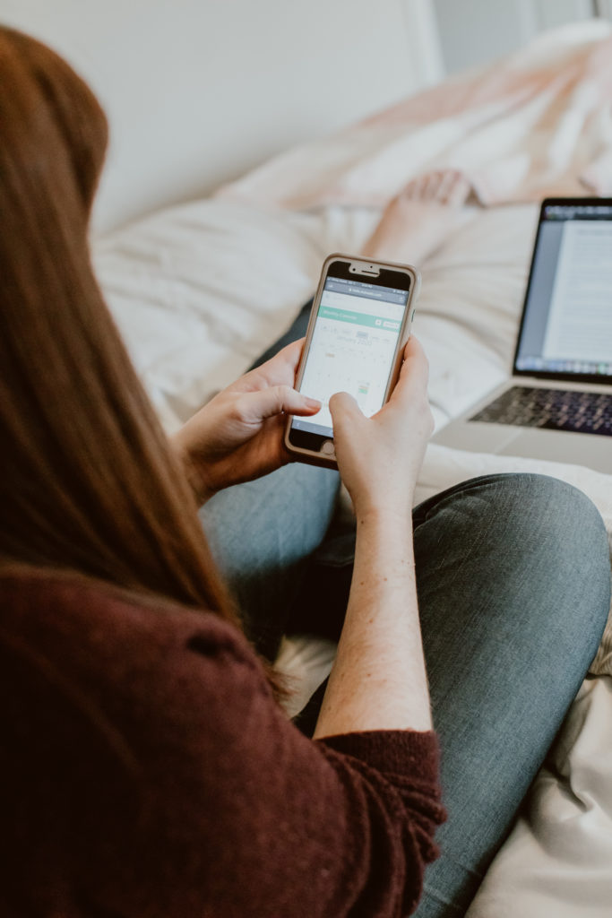 A woman scrolls through a calendar on her phone while sitting in bed. Head to this post for the top 3 reasons to switch from Honeybook to Dubsado.